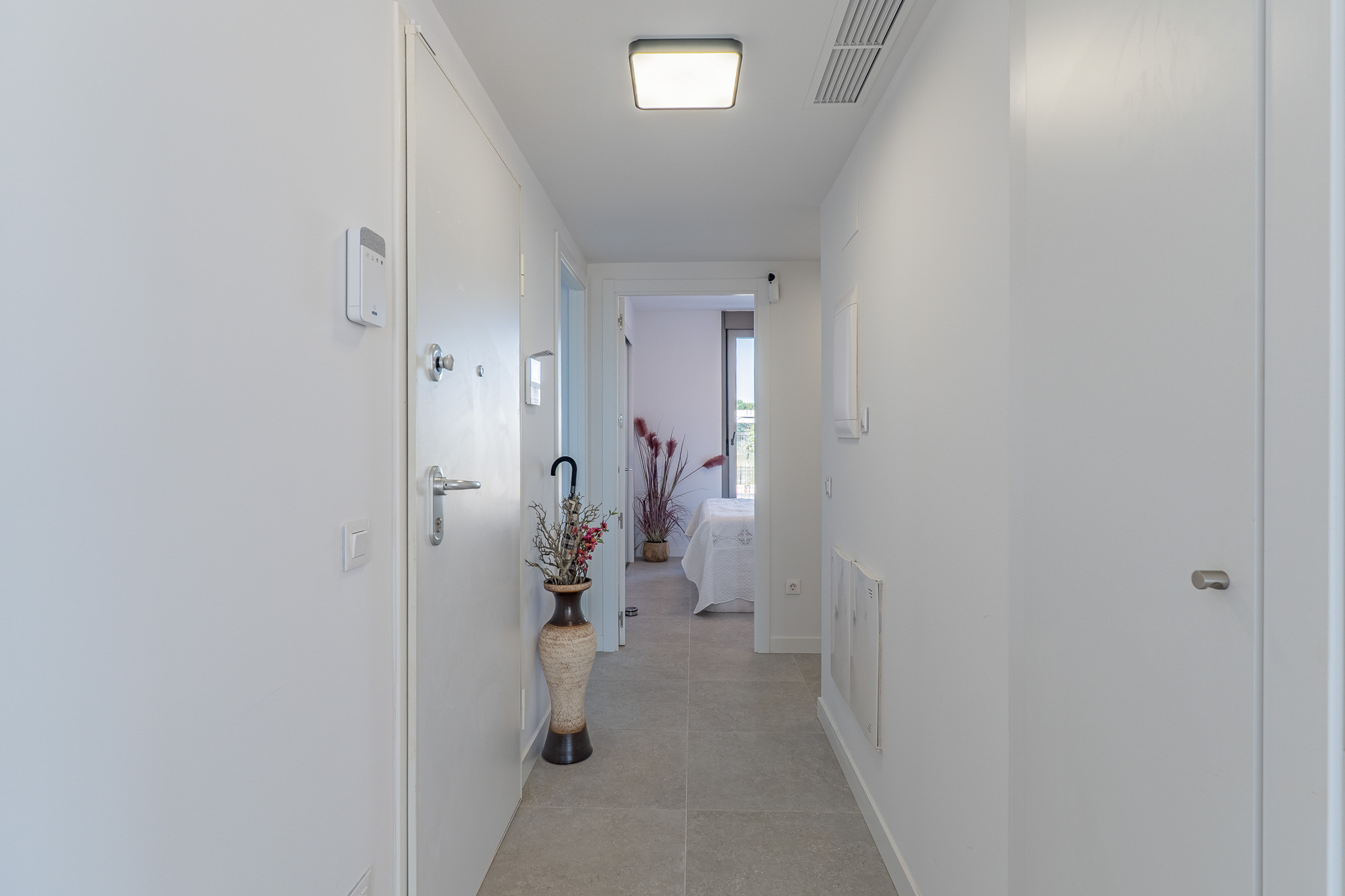 Lichte, moderne hal met lichtgrijze tegels en witte muren. Voorzien van een beveiligingspaneel, eigentijdse plafondverlichting en uitzicht op een slaapkamer.