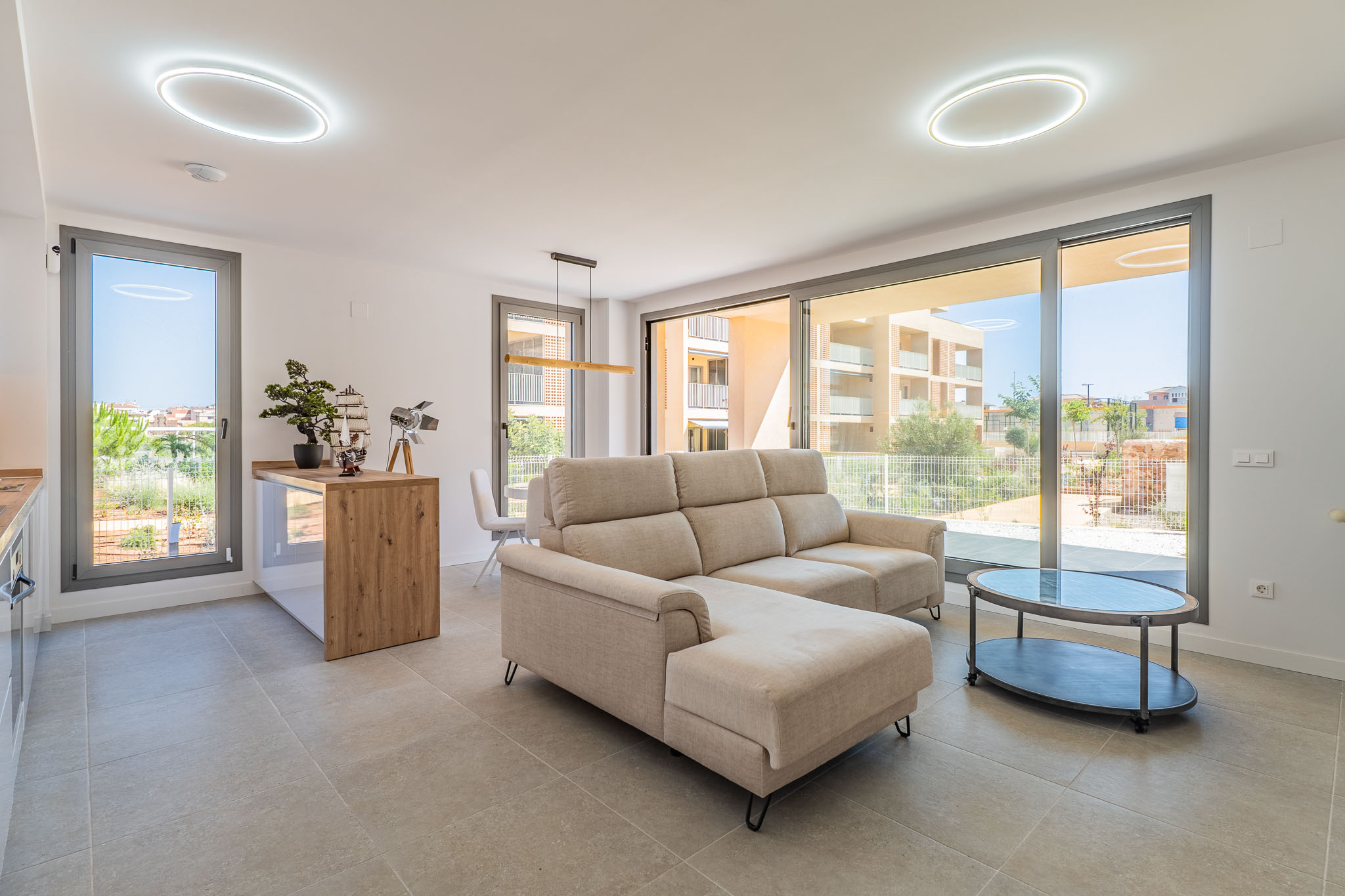 Lichte open woon-eetkamer met beige hoekbank en ovale salontafel. Grote glazen schuifdeuren bieden veel natuurlijk licht en residentieel uitzicht. Open keuken met houten bar.