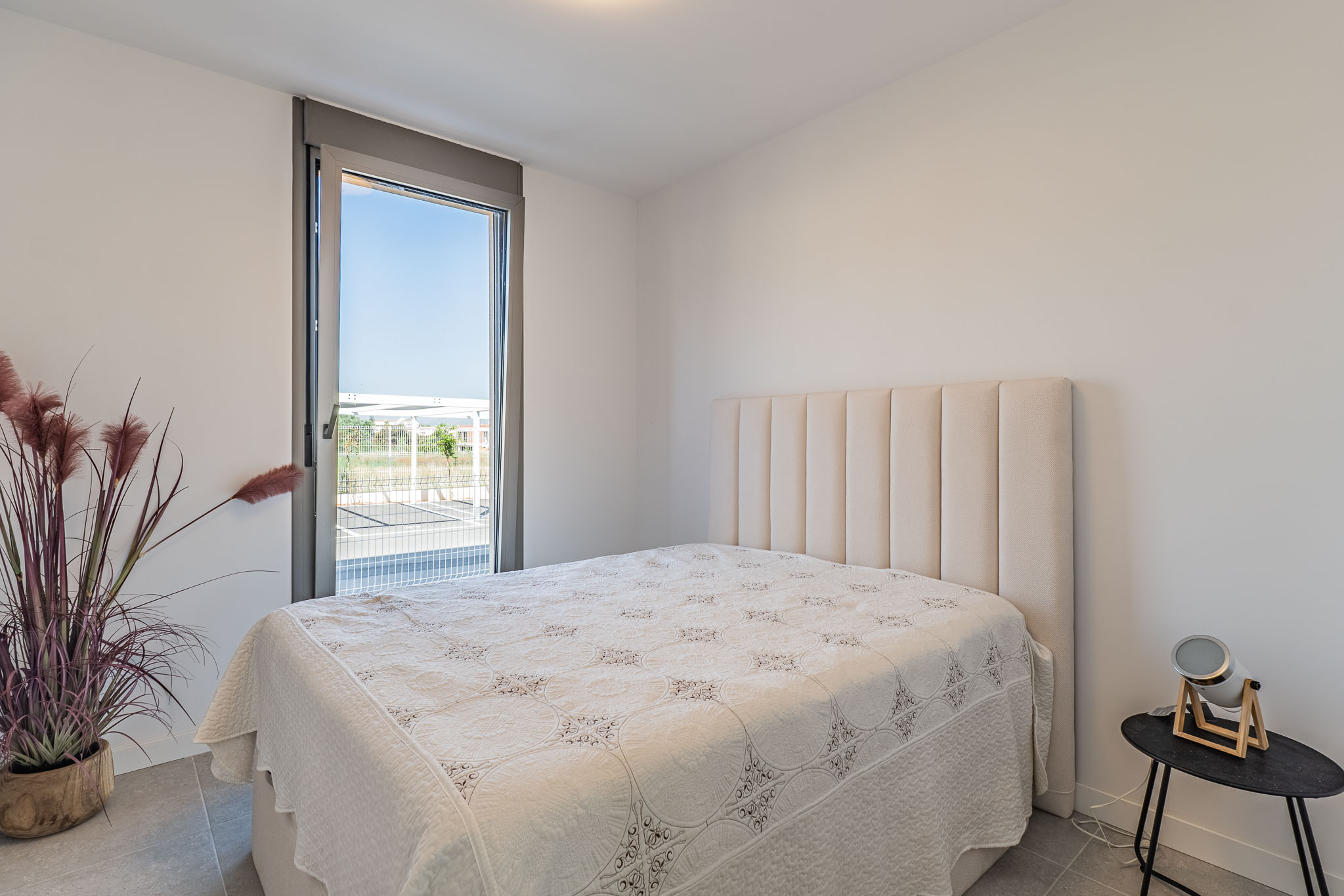 Lichte slaapkamer met bed en hoog, gewatteerd beige hoofdeinde. Groot raam van vloer tot plafond biedt uitzicht naar buiten. Grijze tegelvloer en decoratieve plant.