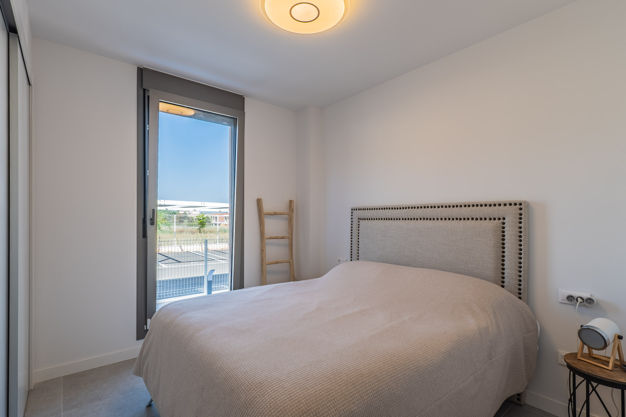 Lichte slaapkamer met bed, gestoffeerd hoofdbord met studs en inbouwkast. Groot raam biedt toegang naar buiten en een grijze tegelvloer.