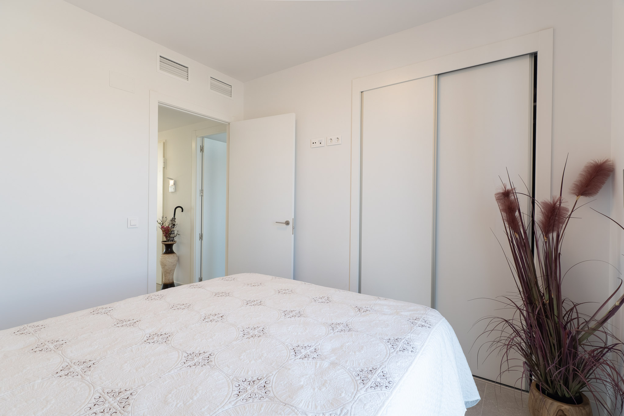 Lichte slaapkamer met bed voorzien van een wit, gedessineerd sprei. Inclusief ingebouwde schuifkast en zichtbare airconditioningroosters in het plafond. Een decoratieve plant verfraait de ruimte.