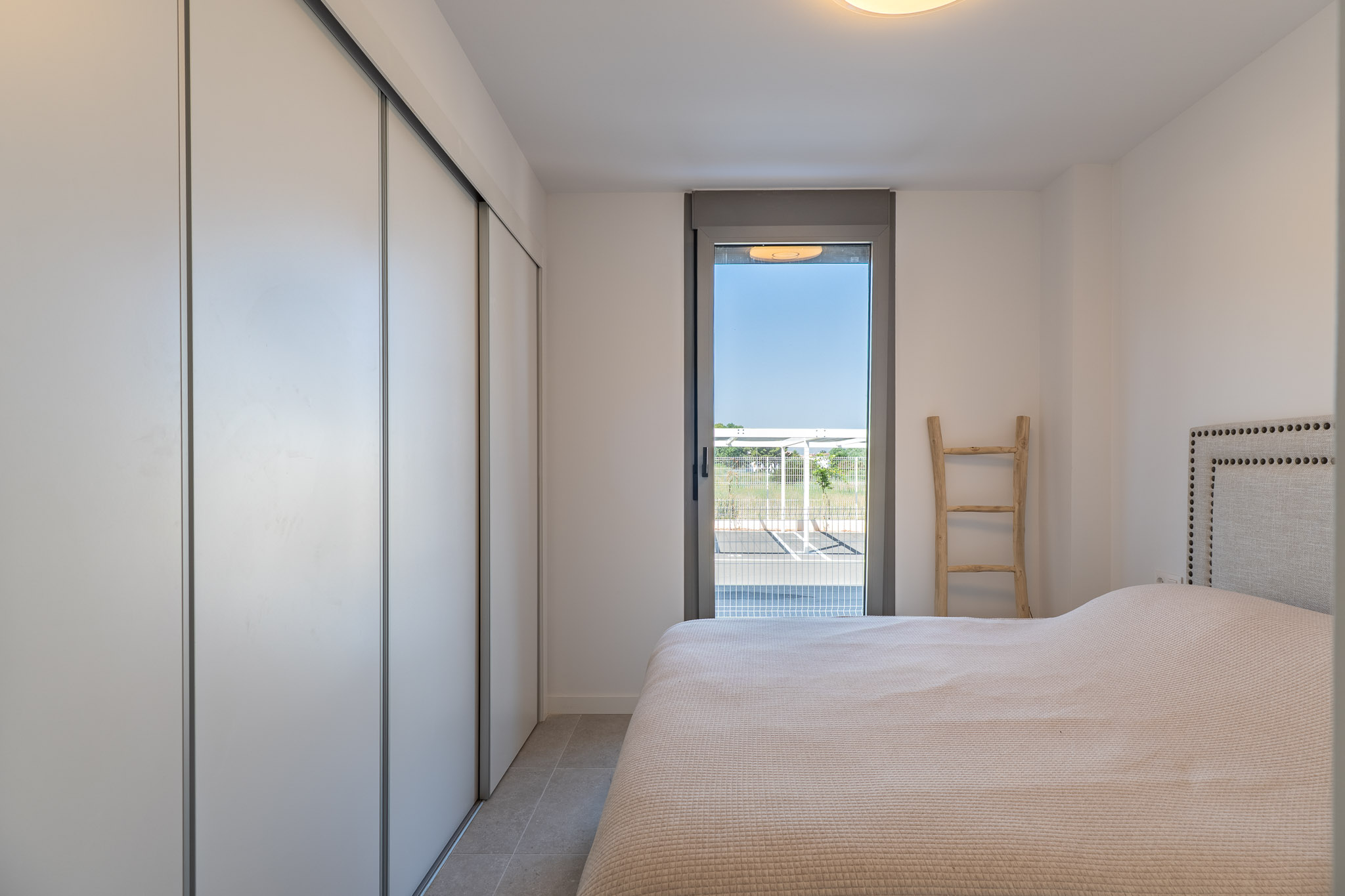 Lichte slaapkamer met ingebouwde kledingkast met witte schuifdeuren, tegelvloer en directe toegang tot de buitenruimte via een glazen deur. Bed met gestoffeerd hoofdbord.