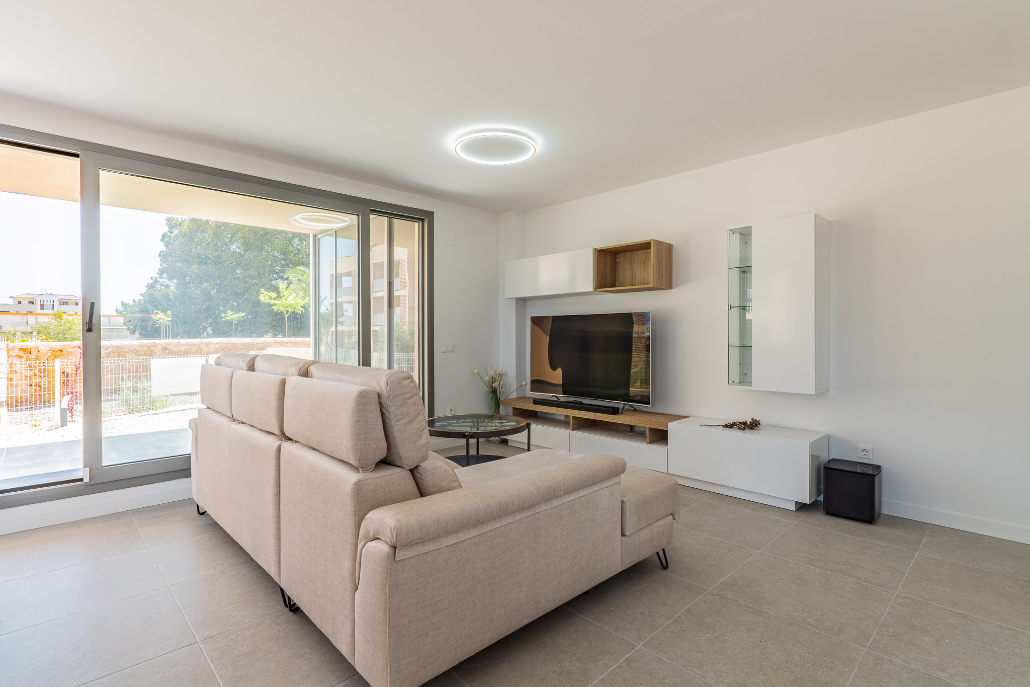 Lichte woonkamer met beige hoekbank, lichtgrijze tegelvloer en grote schuifpuien naar de buitenruimte. Modern tv-meubel met witte en houten opbergelementen.