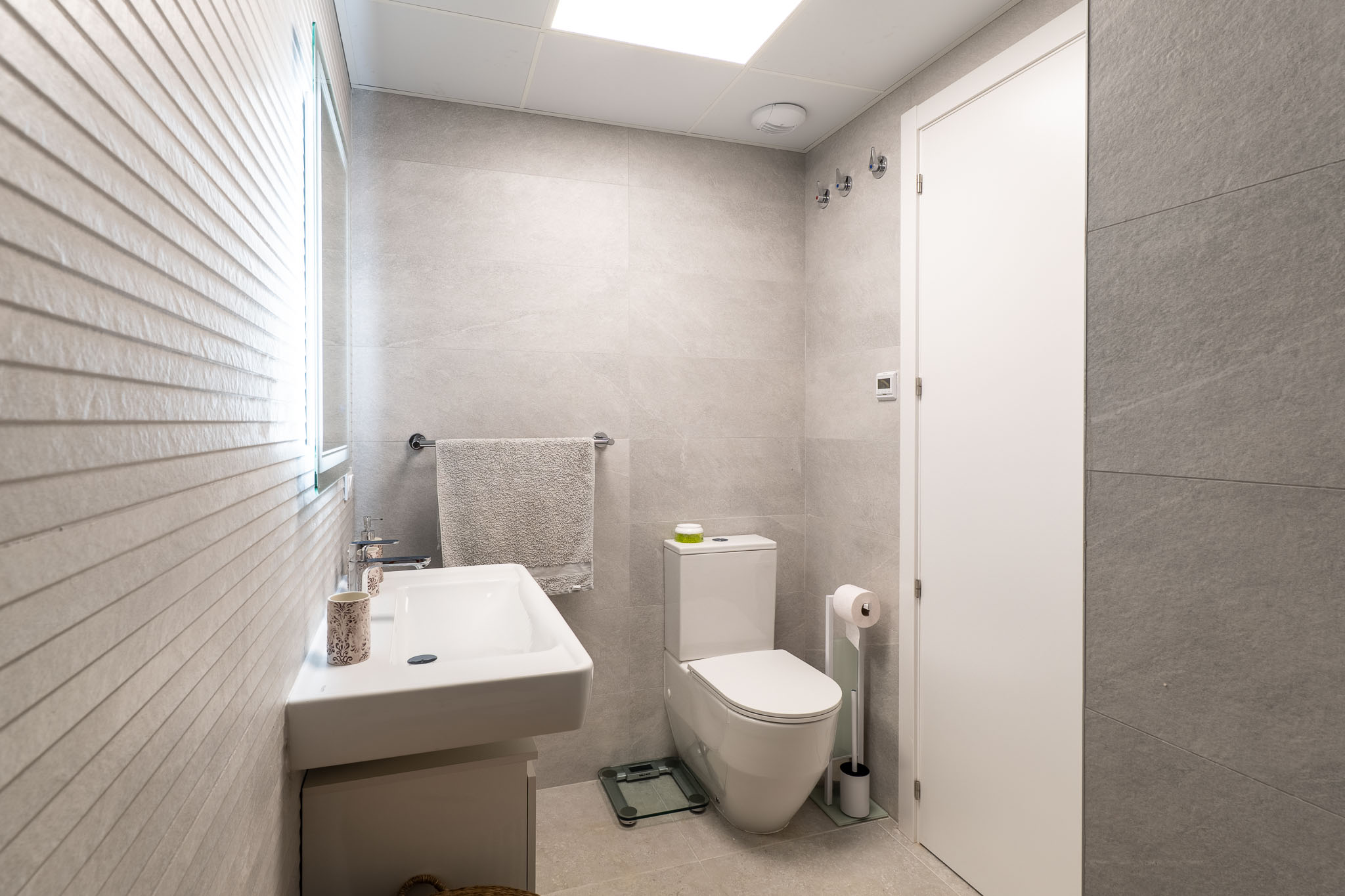 Moderne badkamer met lichtgrijze grootformaat tegels, een wandgemonteerde wastafel en toilet. De verlichte spiegel accentueert de getextureerde muur.