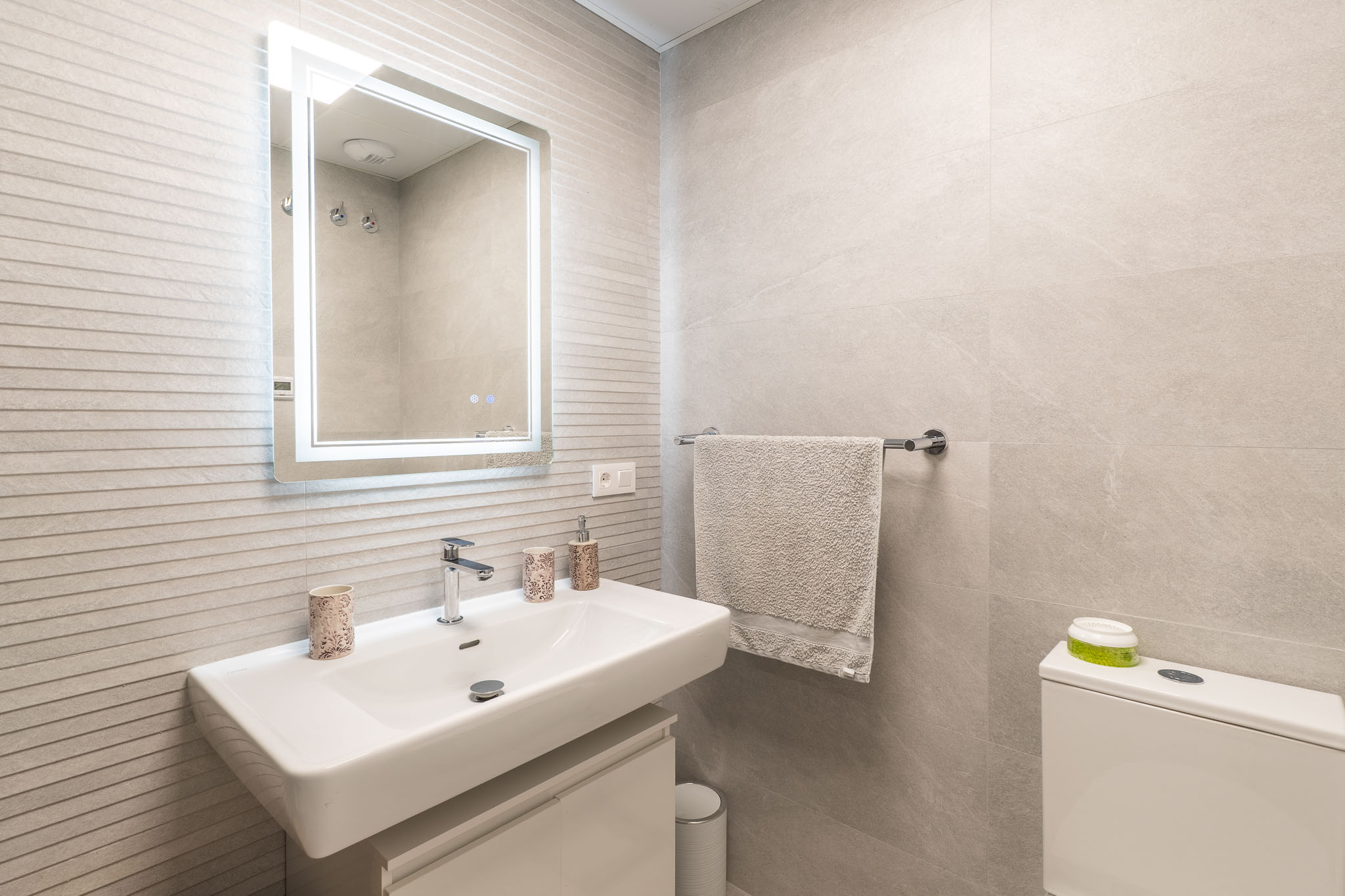 Moderne badkamer met wit rechthoekig wastafelmeubel, wandkraan, verlichte spiegel en lichtgrijze gestructureerde wandtegels.