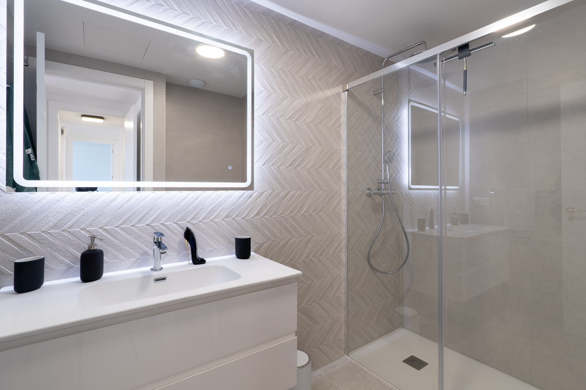 Moderne badkamer met wit wastafelmeubel, verlichte spiegel en wandtegels in visgraatmotief. De glazen douchecabine is voorzien van een regendouche en chromen kranen.