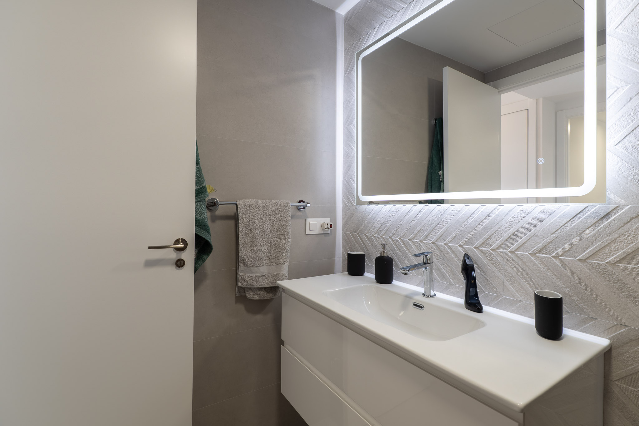 Moderne badkamer met wit zwevend wastafelmeubel, chromen kraan en verlichte spiegel. Gekenmerkt door de getextureerde wandtegels met visgraatmotief.