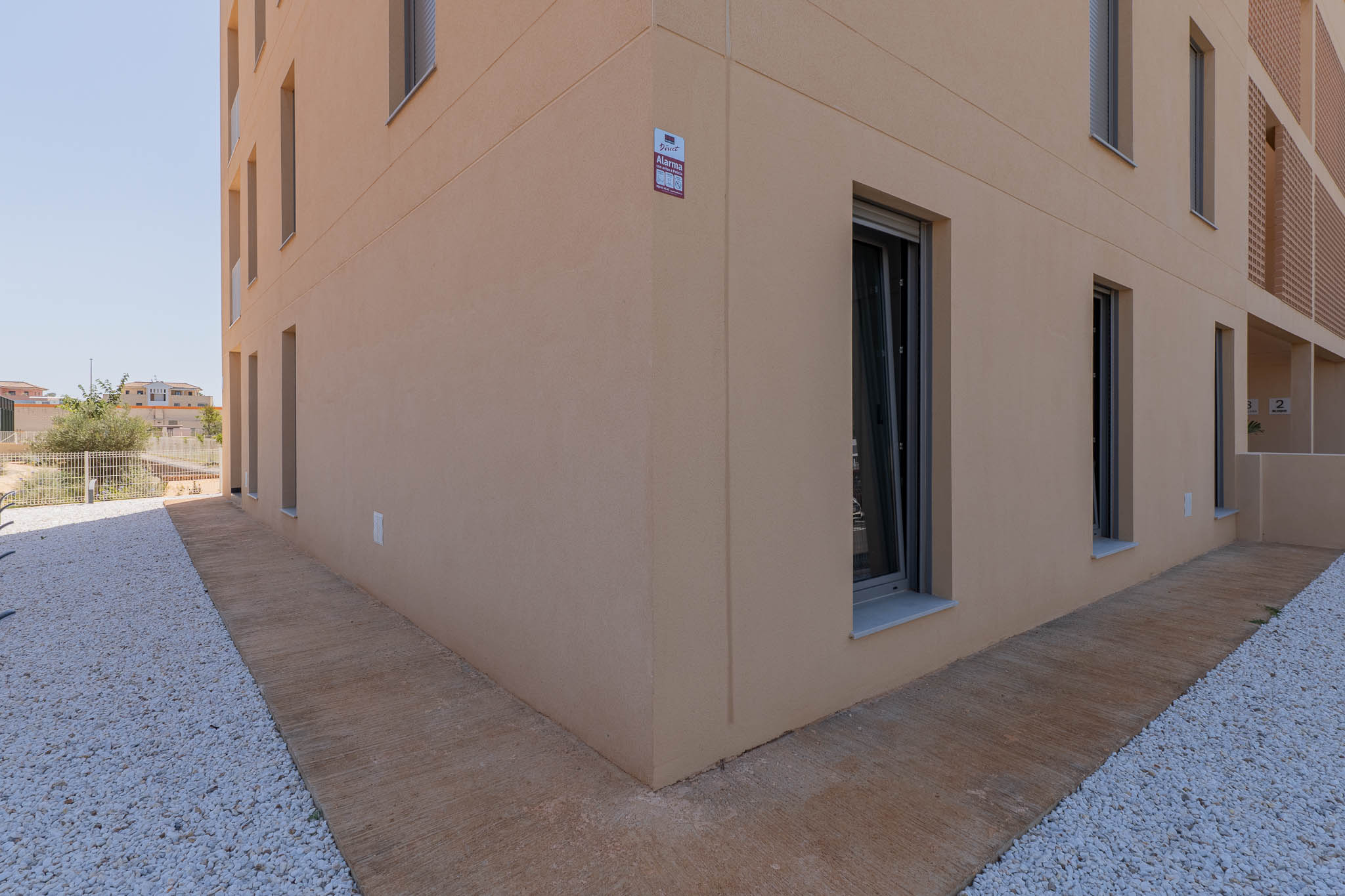 Moderne buitengevel met lichtbeige stucwerk en donker omkaderde ramen. De begane grond heeft een gestructureerd betonnen pad naast een wit grindlandschap, met een zichtbaar alarmsysteem.