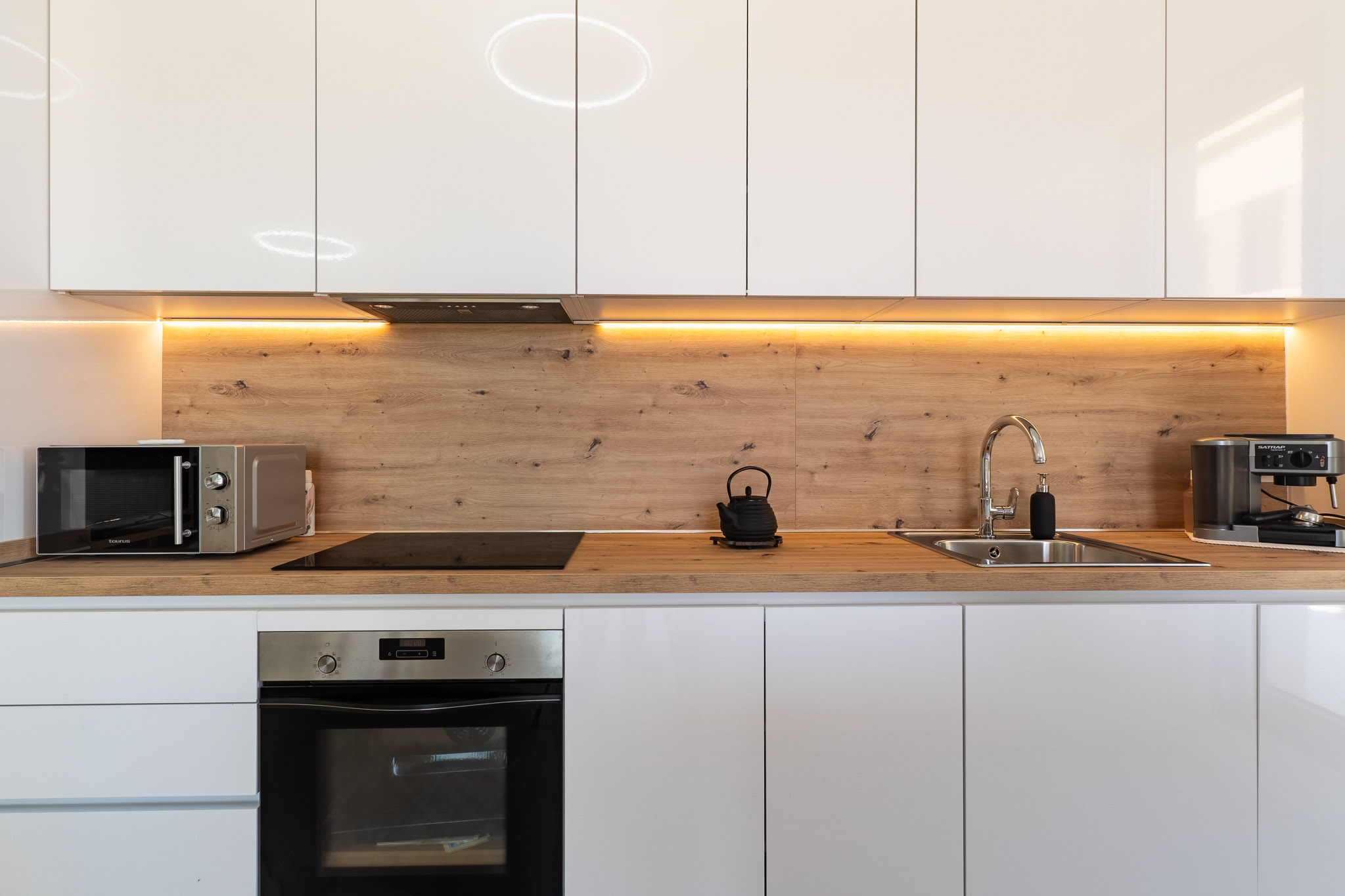 Moderne keuken met glanzend witte kasten, een werkblad en achterwand in houtlook, en warme LED-onderbouwverlichting. Voorzien van geïntegreerde oven, inductiekookplaat en magnetron.