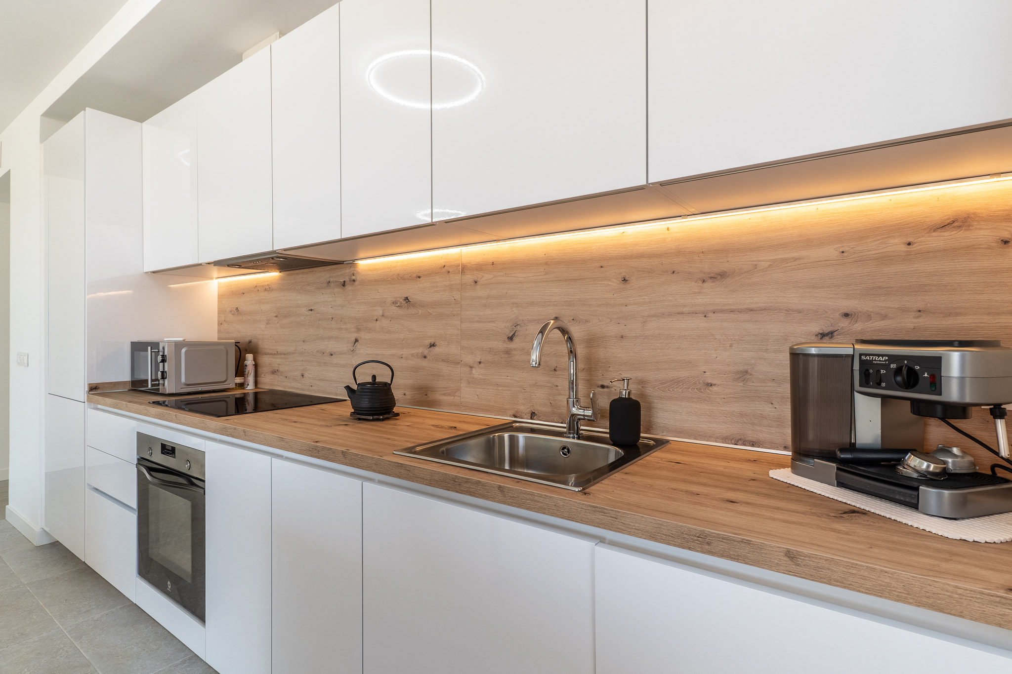 Moderne keuken met glanzend witte kasten, licht houten aanrechtblad en bijpassende achterwand. Voorzien van een geïntegreerde oven, inductiekookplaat en RVS spoelbak, verlicht door LED-onderbouwverlichting.