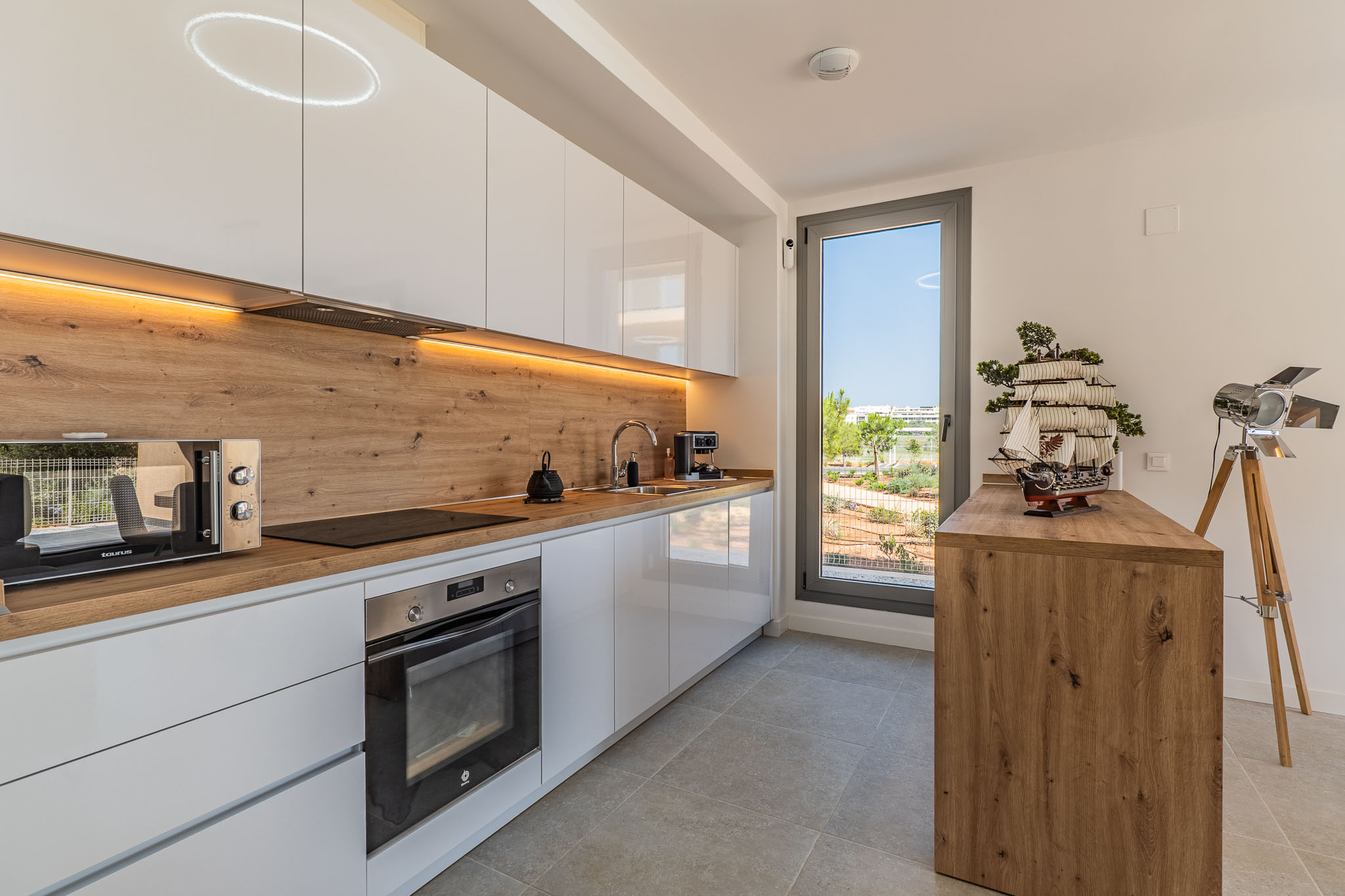 Moderne keuken met hoogglans witte kasten, houtlook werkblad en achterwand. Voorzien van geïntegreerde apparatuur, LED-verlichting en een raam met uitzicht op de tuin.