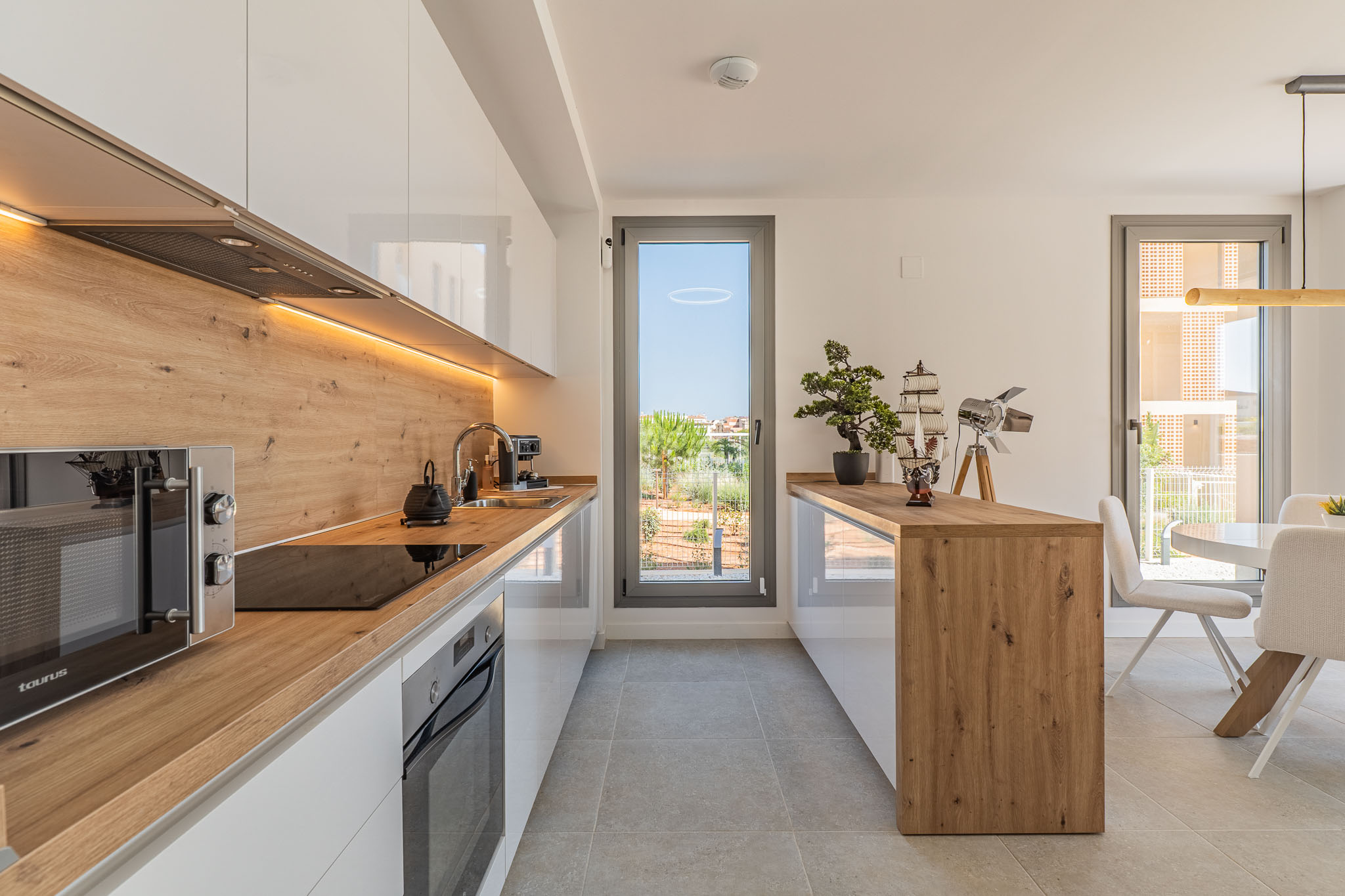 Moderne open keuken met glanzend witte kasten, werkbladen en spatwand in houtlook, en geïntegreerde apparatuur. Voorzien van LED-verlichting onder de bovenkasten en toegang tot buiten.