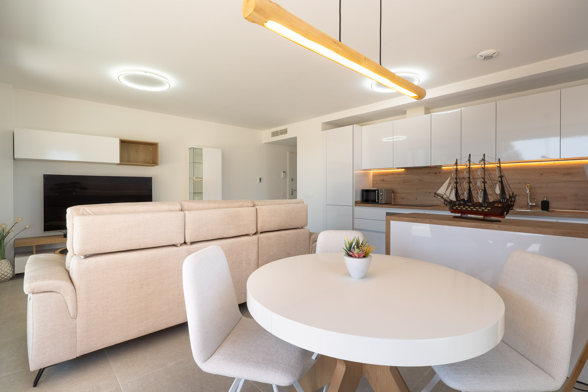 Open woon- en eetkamer met geïntegreerde keuken. Voorzien van een lichte bank, witte ovale eettafel en een moderne keuken met hoogglans witte kasten en houten accenten.