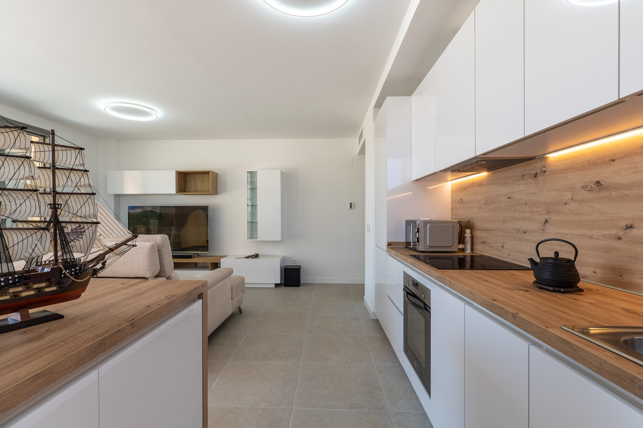 Open woon- en eetruimte met een moderne keuken. De keuken heeft witte hoogglans kasten, licht houtlook werkblad, geïntegreerde apparatuur en warme LED-onderbouwverlichting. Grijze tegelvloer verbindt de ruimtes.