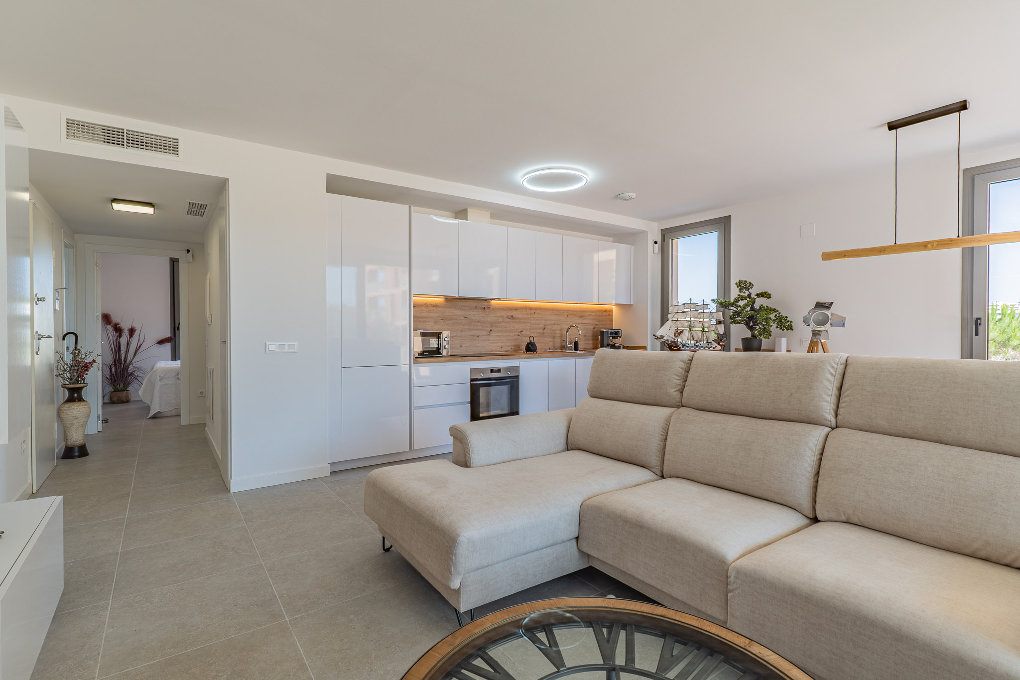 Open woonkamer en keuken met beige hoekbank, moderne witte kasten en een lichte houtlook achterwand. Tegelvloer en veel natuurlijk licht via grote ramen.