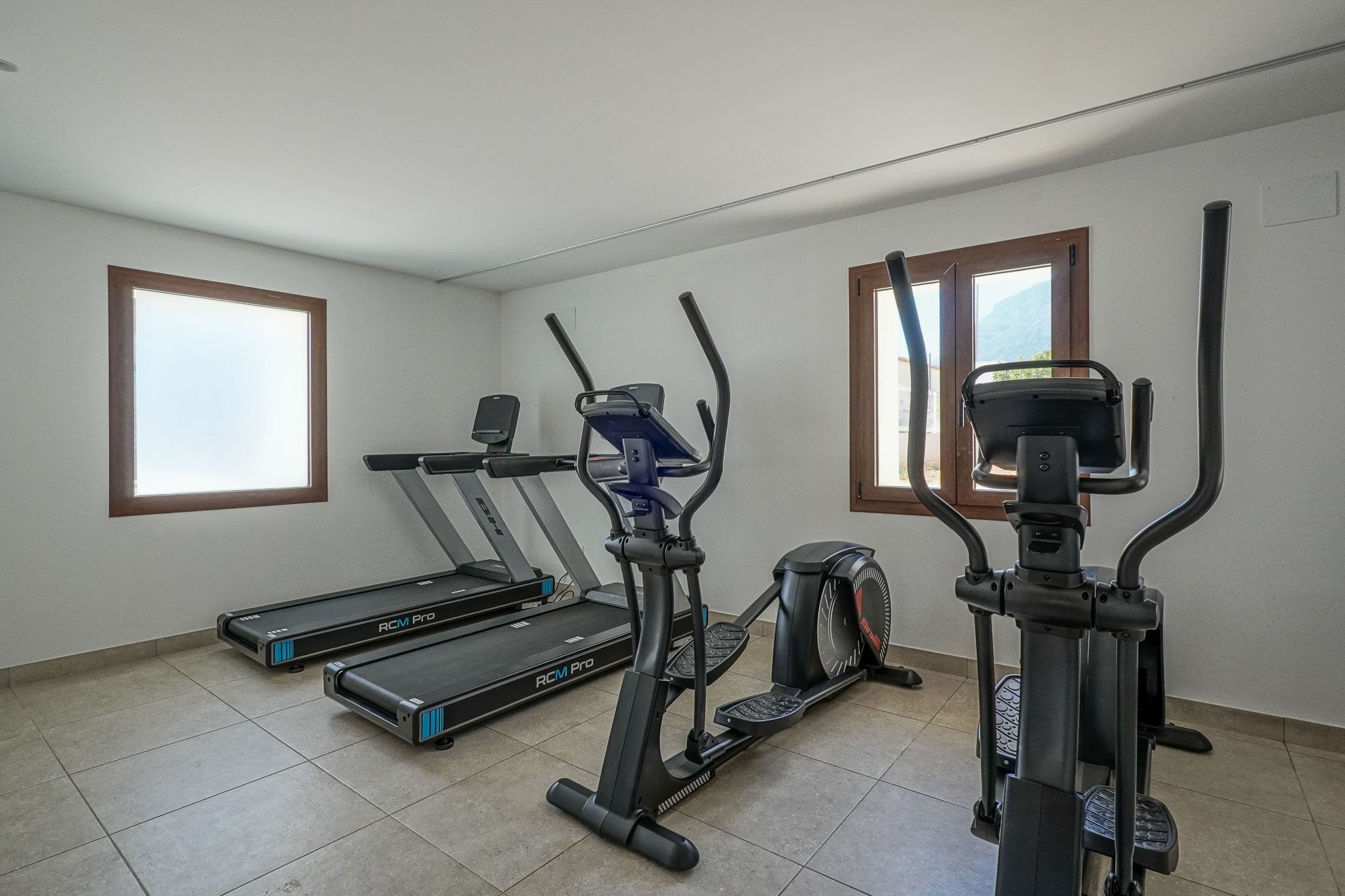 Residentiële fitnessruimte met twee BH RCM Pro loopbanden en twee crosstrainers. Voorzien van een tegelvloer, witte muren en houten raamkozijnen, waarvan één met uitzicht op de bergen.