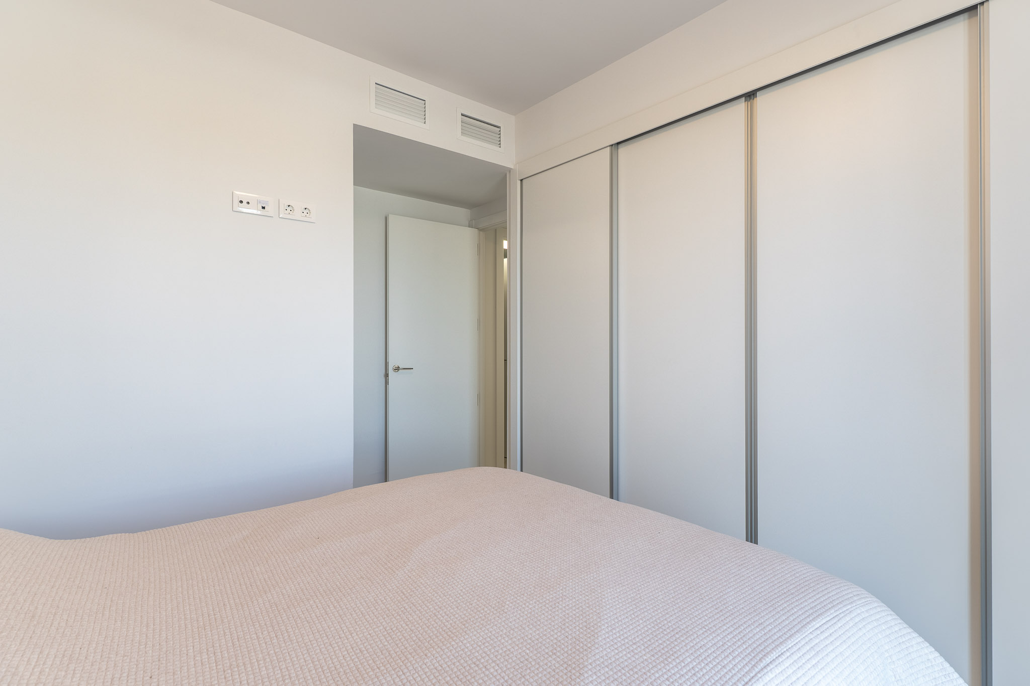 Slaapkamer met een ruime inbouwkast voorzien van witte schuifdeuren. De muur heeft moderne stopcontacten en geïntegreerde ventilatieroosters boven de aangrenzende deur.