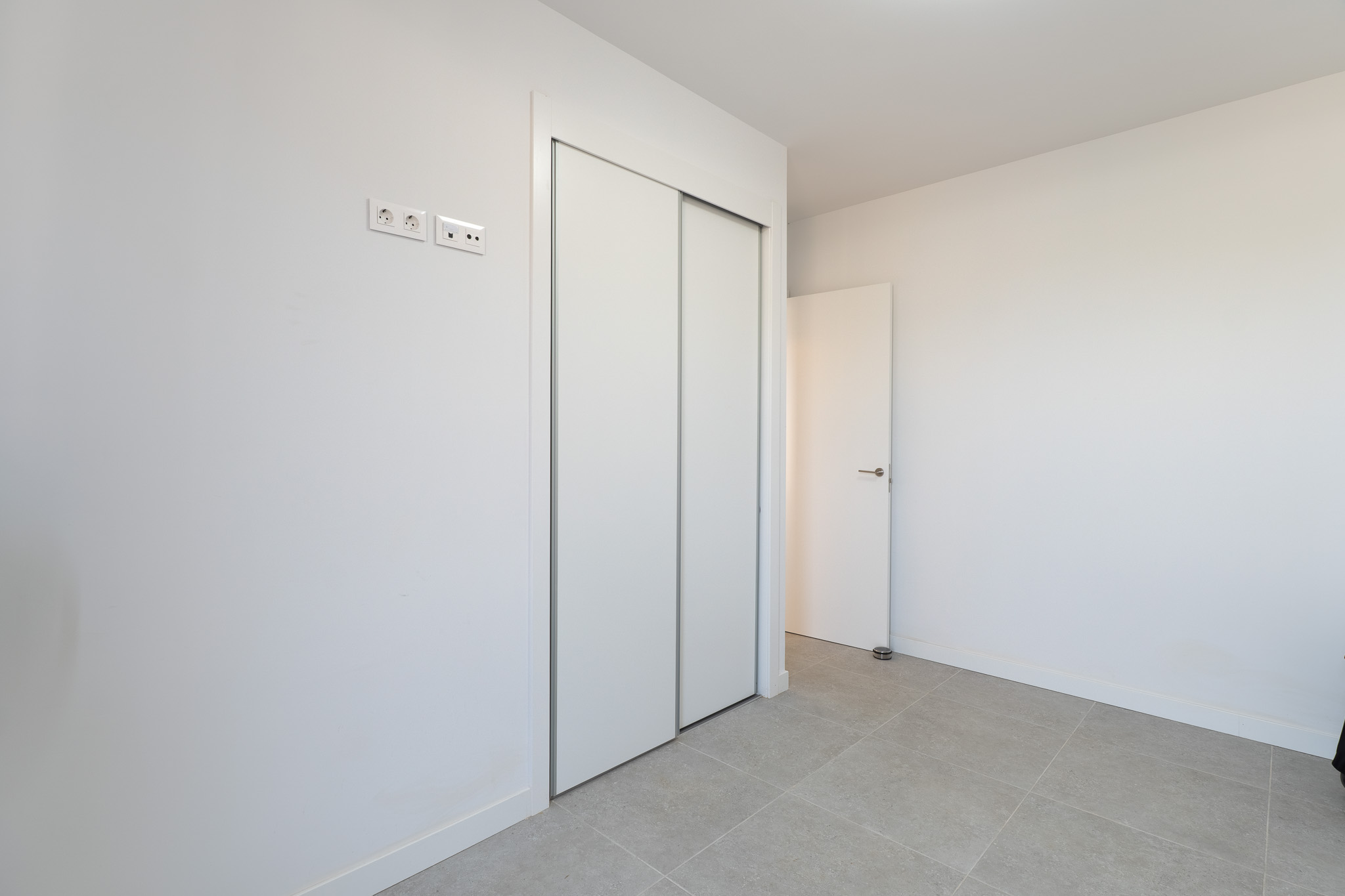 Slaapkamer met ruime inbouwkast voorzien van witte schuifdeuren en een lichtgrijze tegelvloer. Moderne stroom- en data-aansluitingen zijn aanwezig.