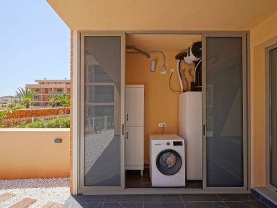 Buitenwasruimte met Bosch wasmachine en aerothermisch systeem, bereikbaar via een betegeld terras met siergrind en tuinzicht.