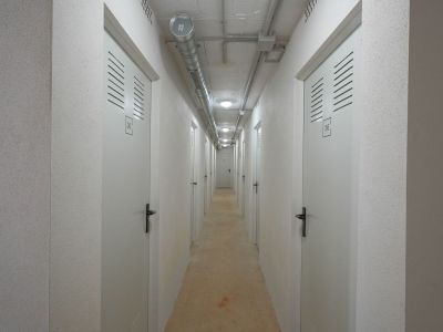Functionele gang naar opslagruimtes, met genummerde witte deuren en ventilatiesleuven. Zichtbetonplafond met industriële leidingen en inbouwverlichting, aangevuld met gestructureerde muren.