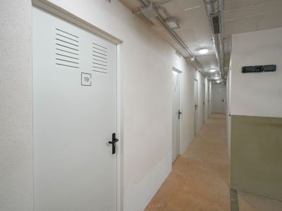 Gang met witte opslagdeuren voorzien van ventilatieroosters en zwarte handgrepen. Het plafond toont zichtbare leidingen en inbouwverlichting boven een betonnen vloer.