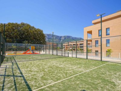 Groene kunstgras padelbaan, omheind en voorzien van verlichting, grenzend aan een kinderspeelplaats en moderne woongebouwen met uitzicht op de bergen.
