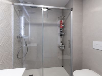 Hedendaagse badkamer met glazen douchecabine en witte douchebak. De douchewand is voorzien van lichtgrijze tegels met een visgraatpatroon, aangevuld met een regendouche en zwevend toilet.