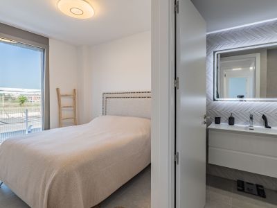 Hoofdslaapkamer met gestoffeerd hoofdbordbed en directe toegang tot een buitenterras. Een en-suite badkamer met zwevend wastafelmeubel en verlichte spiegel is zichtbaar.