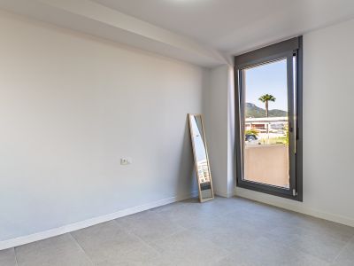 Lege kamer met witte muren en grijze tegelvloer, klaar voor inrichting. Groot raam biedt natuurlijk licht en uitzicht op een palmboom en verre bergen.