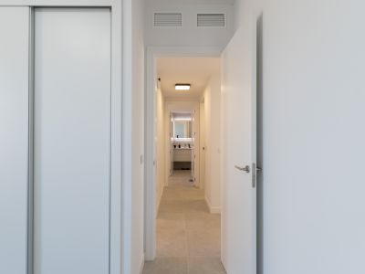 Lichte gang met witte schuifkast en open deur. Lichte vloertegels leiden naar een moderne badkamer met verlichte spiegel en eigentijds badmeubel. Ventilatieopeningen in het plafond zichtbaar.