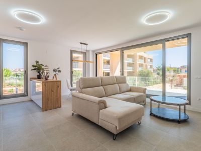 Lichte open woon-eetkamer met beige hoekbank en ovale salontafel. Grote glazen schuifdeuren bieden veel natuurlijk licht en residentieel uitzicht. Open keuken met houten bar.