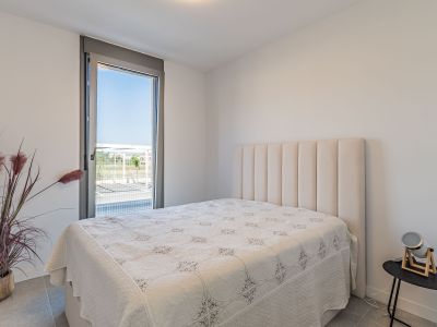 Lichte slaapkamer met bed en hoog, gewatteerd beige hoofdeinde. Groot raam van vloer tot plafond biedt uitzicht naar buiten. Grijze tegelvloer en decoratieve plant.