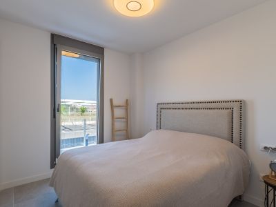 Lichte slaapkamer met bed, gestoffeerd hoofdbord met studs en inbouwkast. Groot raam biedt toegang naar buiten en een grijze tegelvloer.