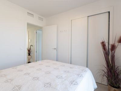 Lichte slaapkamer met bed voorzien van een wit, gedessineerd sprei. Inclusief ingebouwde schuifkast en zichtbare airconditioningroosters in het plafond. Een decoratieve plant verfraait de ruimte.