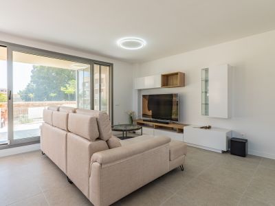 Lichte woonkamer met beige hoekbank, lichtgrijze tegelvloer en grote schuifpuien naar de buitenruimte. Modern tv-meubel met witte en houten opbergelementen.