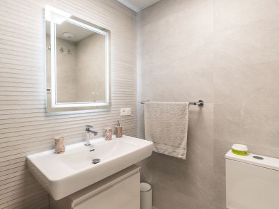 Moderne badkamer met wit rechthoekig wastafelmeubel, wandkraan, verlichte spiegel en lichtgrijze gestructureerde wandtegels.