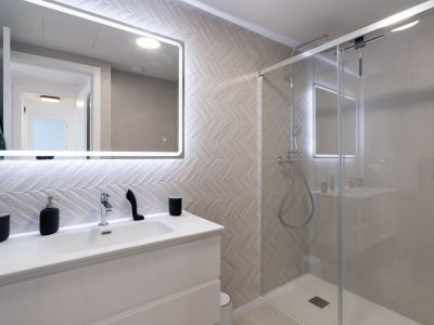 Moderne badkamer met wit wastafelmeubel, verlichte spiegel en wandtegels in visgraatmotief. De glazen douchecabine is voorzien van een regendouche en chromen kranen.
