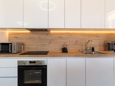 Moderne keuken met glanzend witte kasten, een werkblad en achterwand in houtlook, en warme LED-onderbouwverlichting. Voorzien van geïntegreerde oven, inductiekookplaat en magnetron.