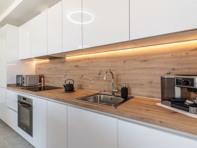 Moderne keuken met glanzend witte kasten, licht houten aanrechtblad en bijpassende achterwand. Voorzien van een geïntegreerde oven, inductiekookplaat en RVS spoelbak, verlicht door LED-onderbouwverlichting.