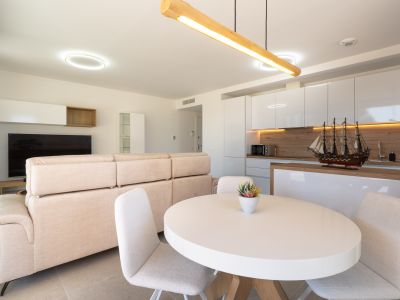 Open woon- en eetkamer met geïntegreerde keuken. Voorzien van een lichte bank, witte ovale eettafel en een moderne keuken met hoogglans witte kasten en houten accenten.