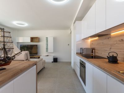 Open woon- en eetruimte met een moderne keuken. De keuken heeft witte hoogglans kasten, licht houtlook werkblad, geïntegreerde apparatuur en warme LED-onderbouwverlichting. Grijze tegelvloer verbindt de ruimtes.