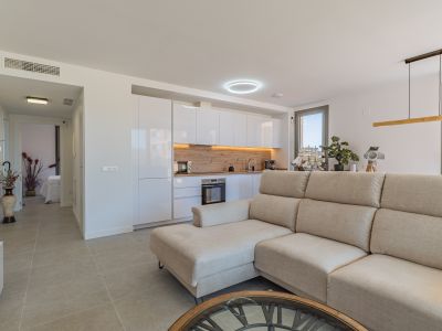 Open woonkamer en keuken met beige hoekbank, moderne witte kasten en een lichte houtlook achterwand. Tegelvloer en veel natuurlijk licht via grote ramen.