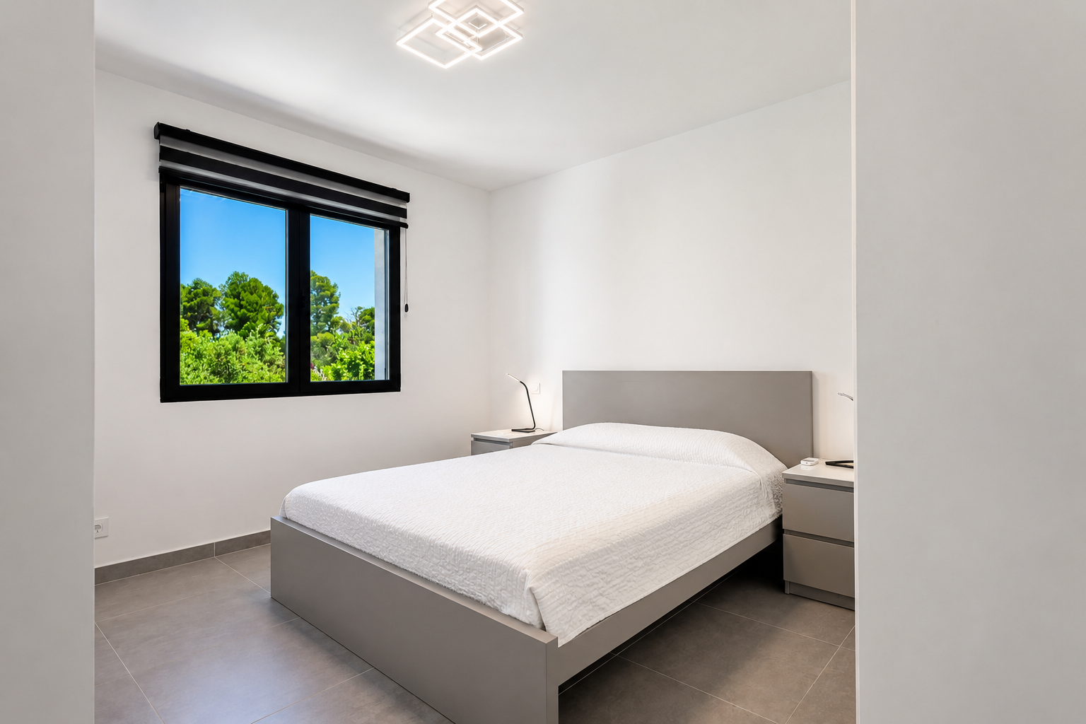 Eigentijdse slaapkamer met grijs platformbed, tegelvloer en natuurlijk licht via een raam met uitzicht op weelderige bomen. Voorzien van moderne plafond- en nachtlampjes.