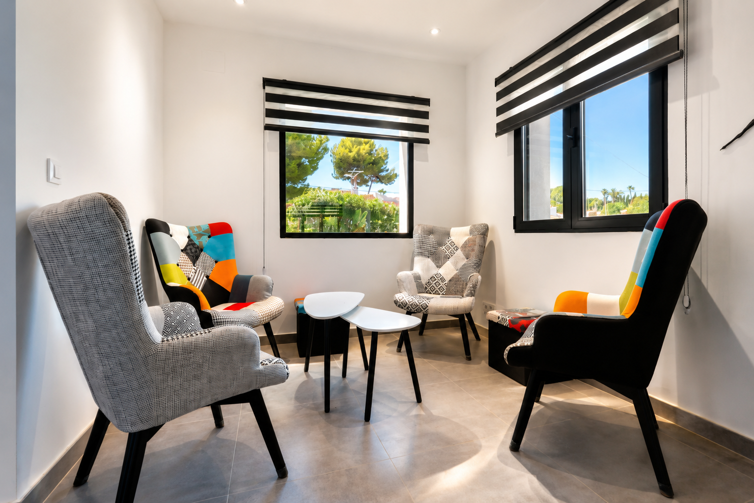 Gezellige zithoek met vier eclectische fauteuils en minimalistische bijzettafels. Ramen met gestreepte jaloezieën bieden natuurlijk licht en uitzicht op het groen.