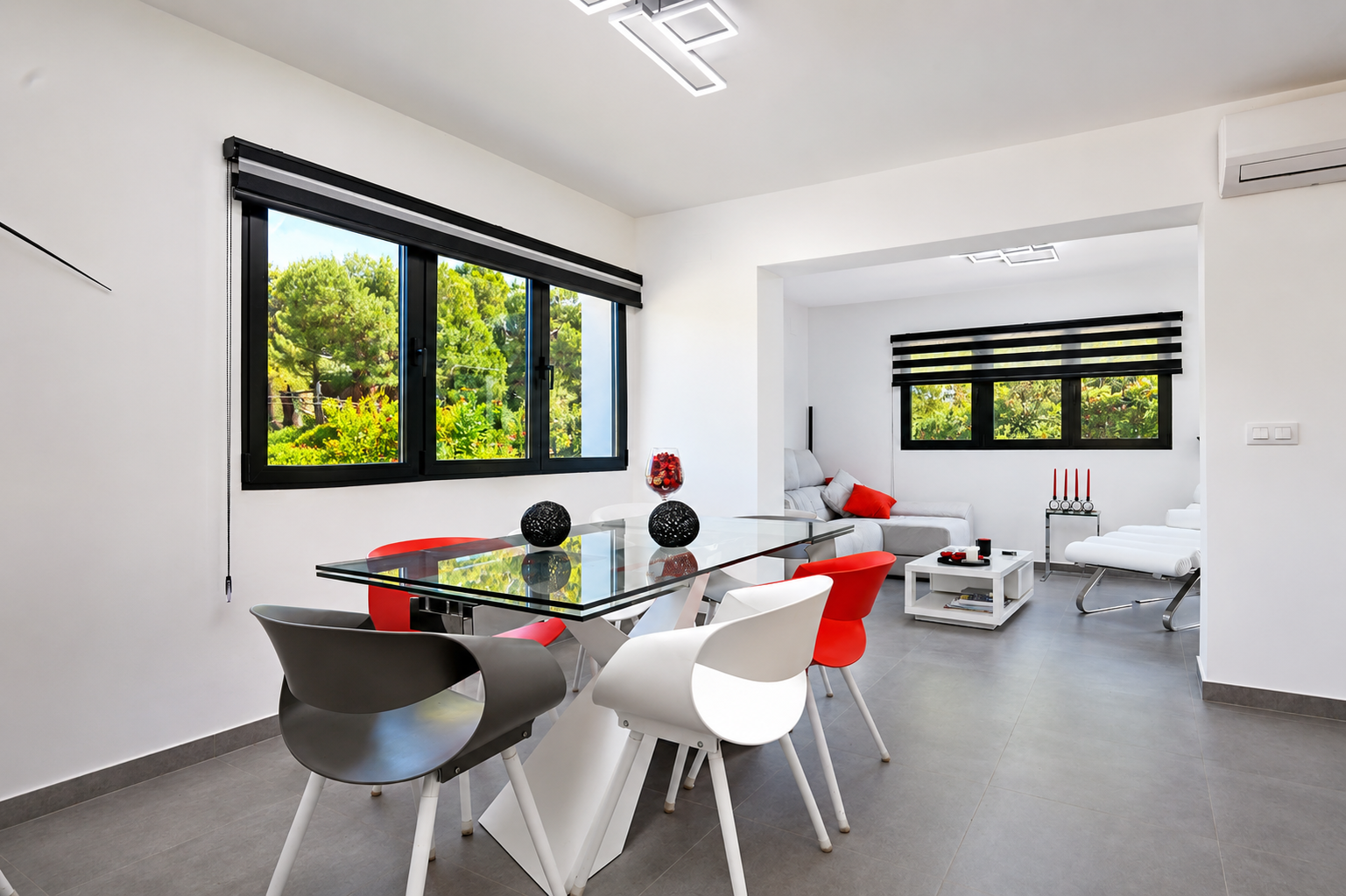 Hedendaagse eetruimte met glazen tafel en designstoelen in wit, rood en grijs. Grote zwart omkaderde ramen bieden uitzicht op de weelderige groene omgeving.