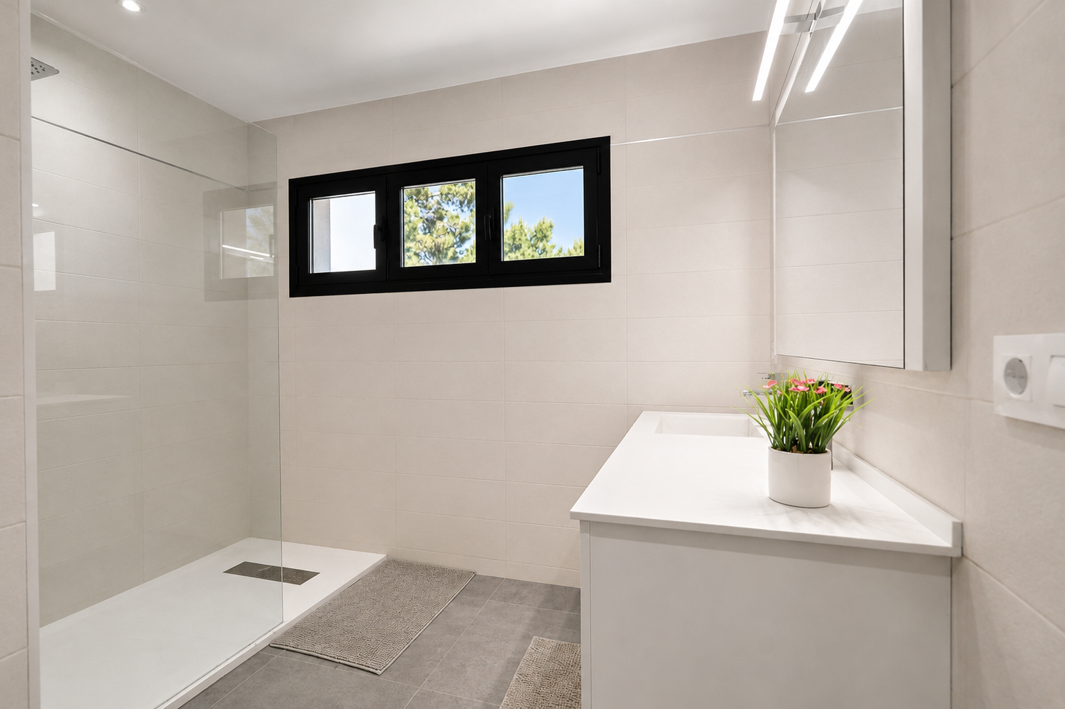 Moderne badkamer met inloopdouche en glazen wand. Lichte tegels, zwart kozijnraam met uitzicht op groen. Wit badmeubel met geïntegreerde wastafel en verlichte spiegel.