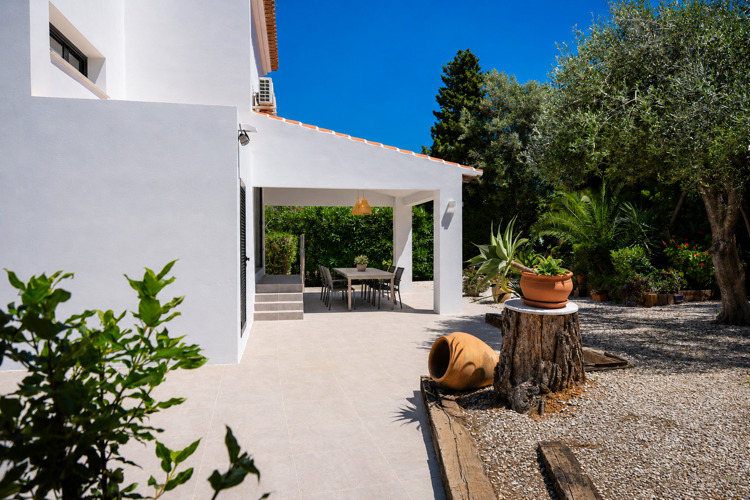 Ruime naya met lichte tegelvloer en eethoek buiten, geflankeerd door een mediterrane tuin met olijfboom en decoratieve elementen.