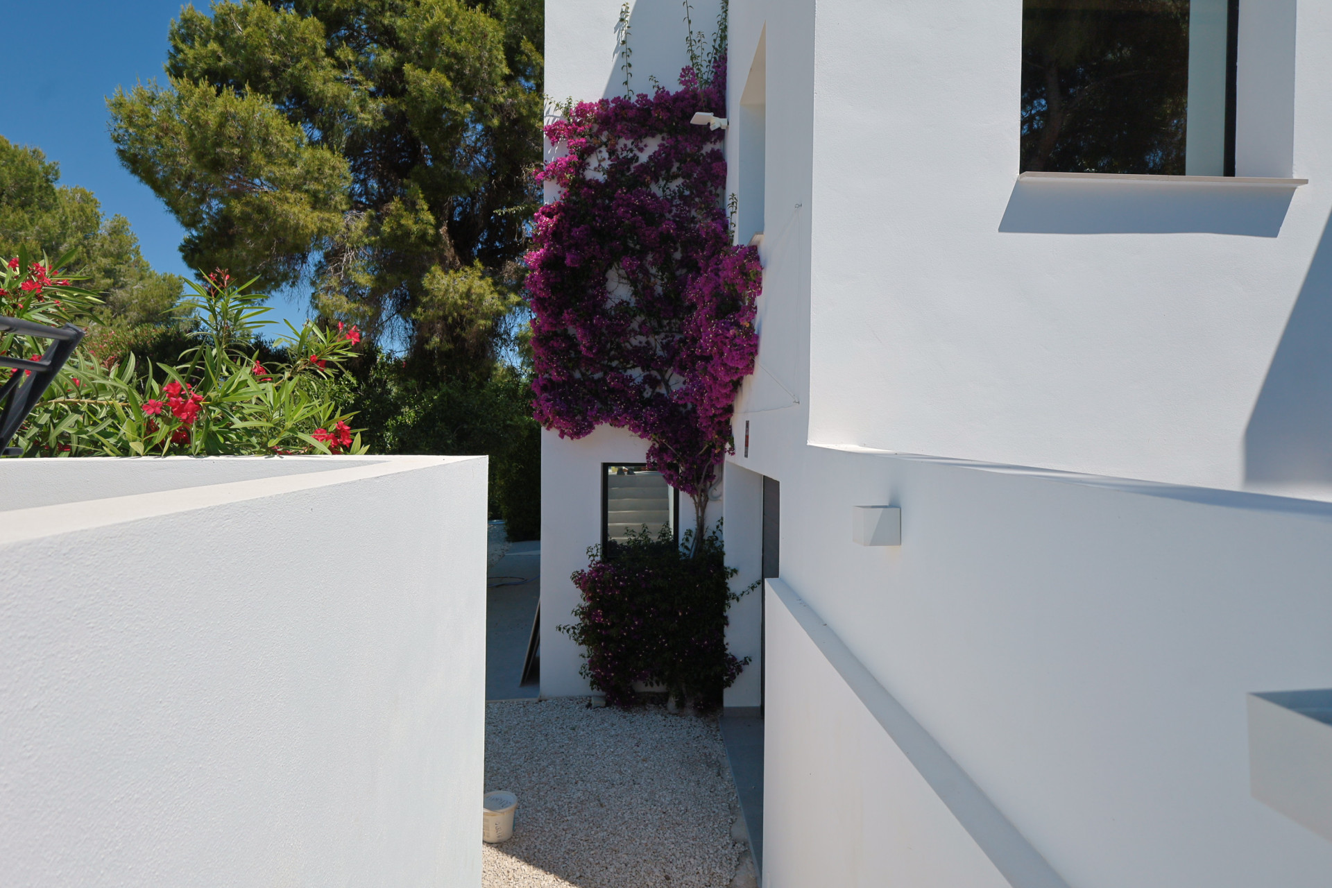 Zonnige doorgang tussen witte muren, versierd met levendige paarse bougainvillea en grindpad. Mediterrane bomen en rode oleanderbloemen omlijsten de helderblauwe lucht.