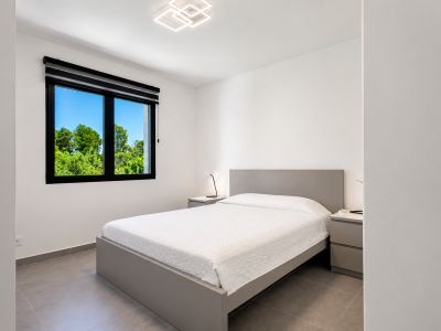 Eigentijdse slaapkamer met grijs platformbed, tegelvloer en natuurlijk licht via een raam met uitzicht op weelderige bomen. Voorzien van moderne plafond- en nachtlampjes.