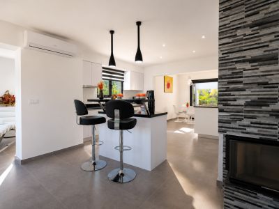 Hedendaagse open keuken met zwart aanrechtblad eiland en barkrukken, witte kasten en inbouwverlichting. De aangrenzende eethoek en een open haard met gestructureerde steenbekleding zijn zichtbaar.
